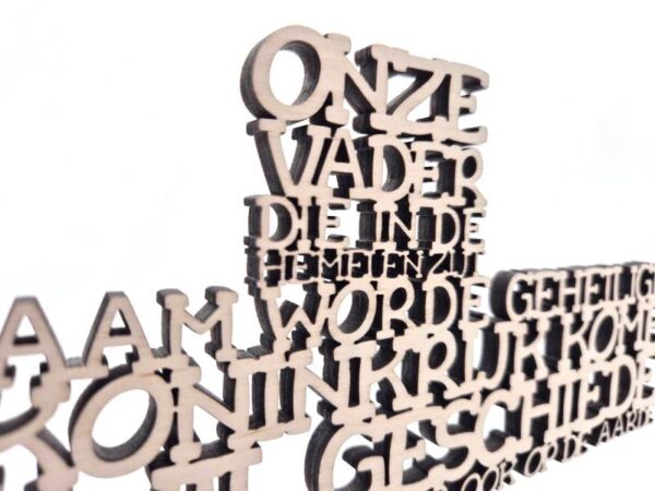Close‑up van een Onze Vader kruis met uitgesneden Nederlandse tekst als moderne typografische geloofsdecoratie.